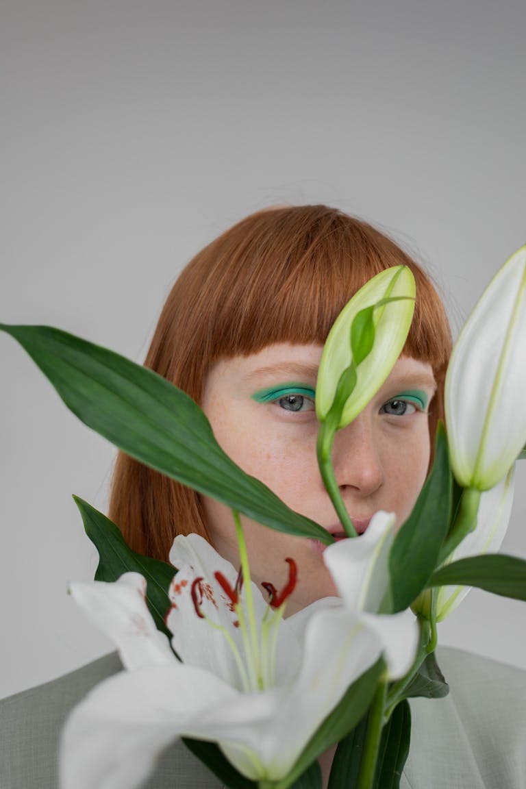 Portrait of a redheaded woman partially concealed by white lilies, showcasing makeup artistry.